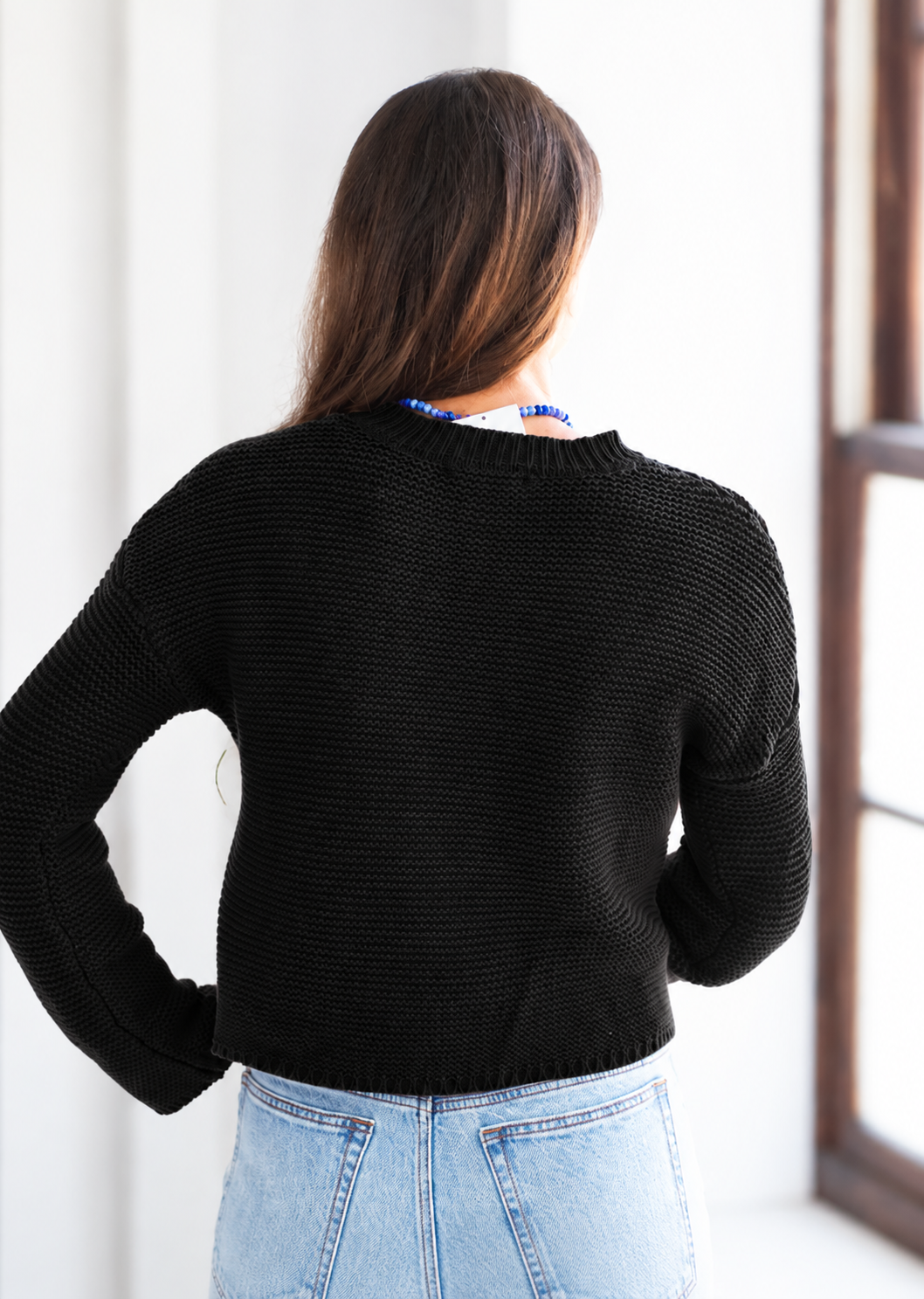 A woman viewed from behind wearing a black textured knit sweater and light blue jeans in a bright indoor setting.