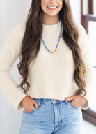 A smiling woman with long wavy brown hair wearing a cream-colored textured knit sweater and light blue jeans, standing indoors near a window.