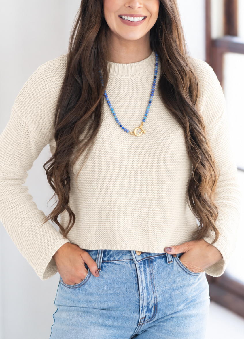 A smiling woman with long wavy brown hair wearing a cream-colored textured knit sweater and light blue jeans, standing indoors near a window.