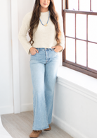 A woman stands in a bright room next to a window, wearing a cream long-sleeve knit top, light-wash blue flare jeans, and brown shoes.