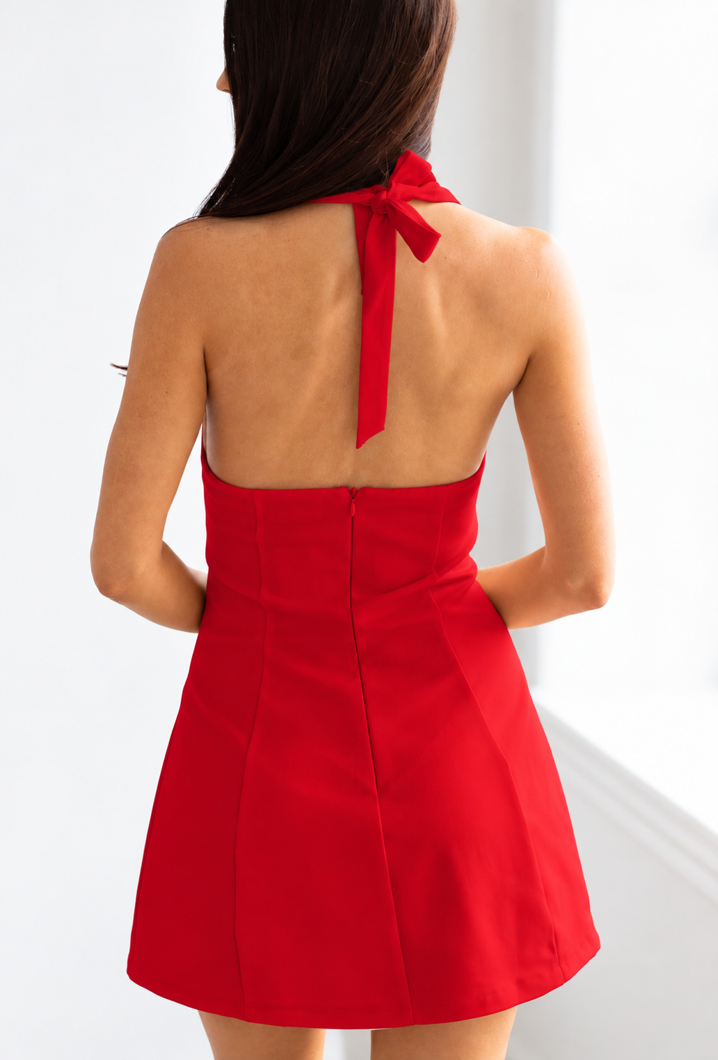 A woman shown from the back wearing a bright red halter-neck mini dress with a tie at the back of the neck and a fitted flared silhouette.