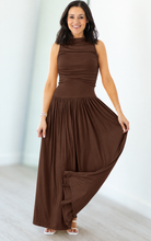 A woman modeling a sleeveless, high-neck, floor-length brown maxi dress with a gathered, draped bodice and flowing skirt, standing in a bright indoor setting.