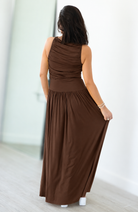 A woman viewed from behind wearing a sleeveless, draped maxi dress in a solid dark brown color, standing in a brightly lit indoor space with light-colored walls and flooring.