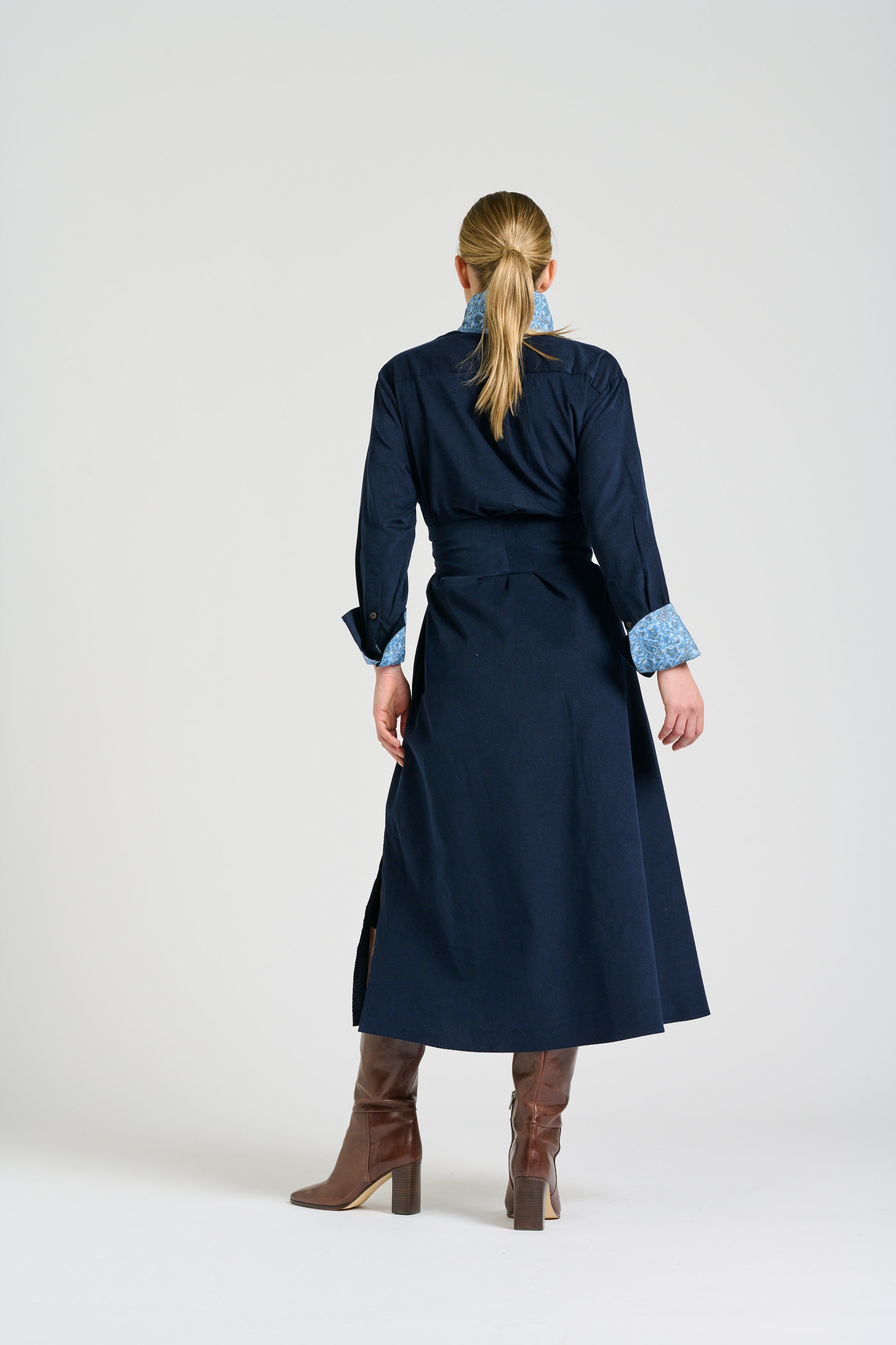 A woman viewed from the back wearing a long navy wrap-style coat with light blue patterned cuffs, brown knee-high boots, and a ponytail, photographed against a plain light gray studio background.