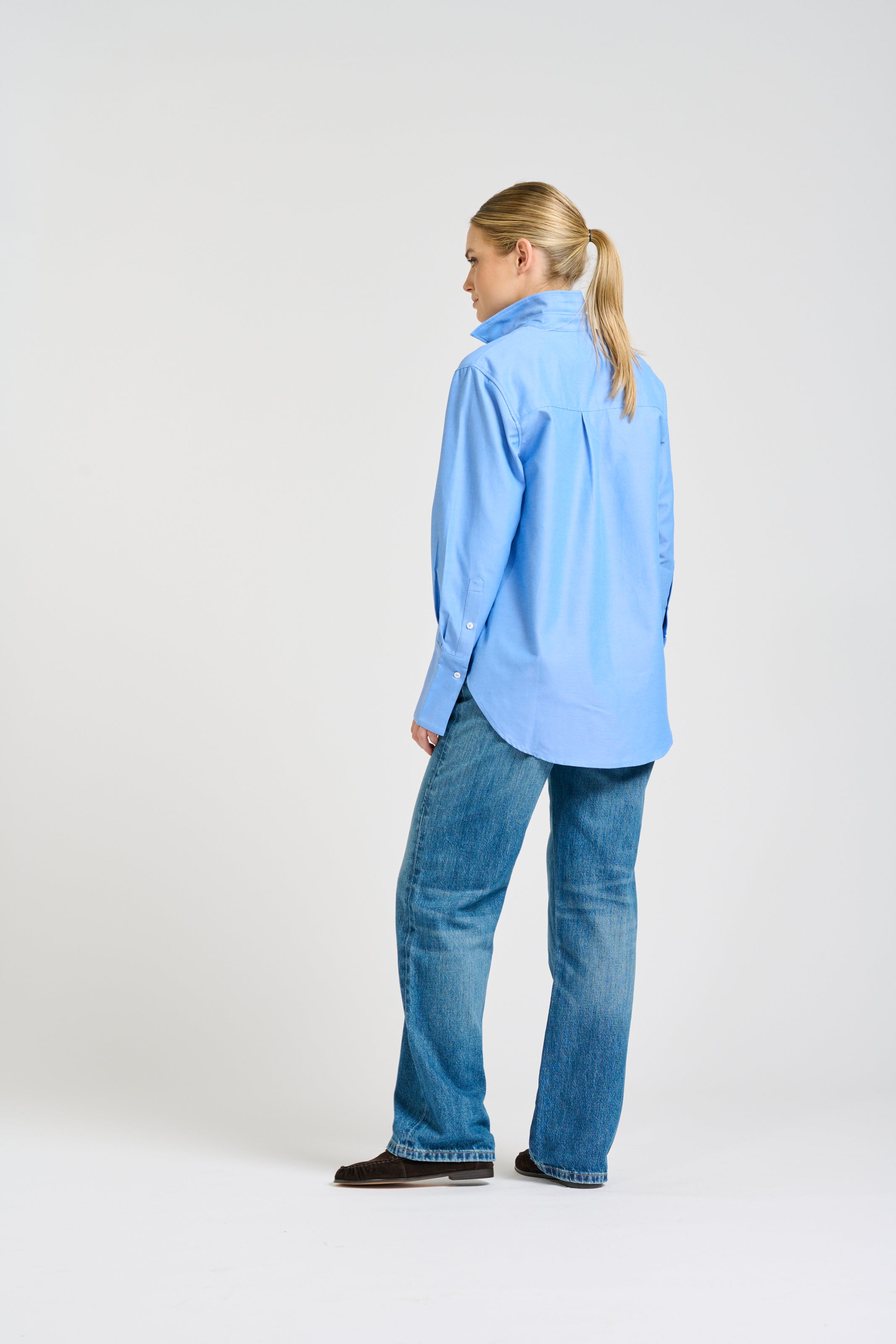 A woman with blonde hair in a ponytail is shown from the back wearing a light blue button-down shirt and blue jeans, standing against a plain light gray studio background.