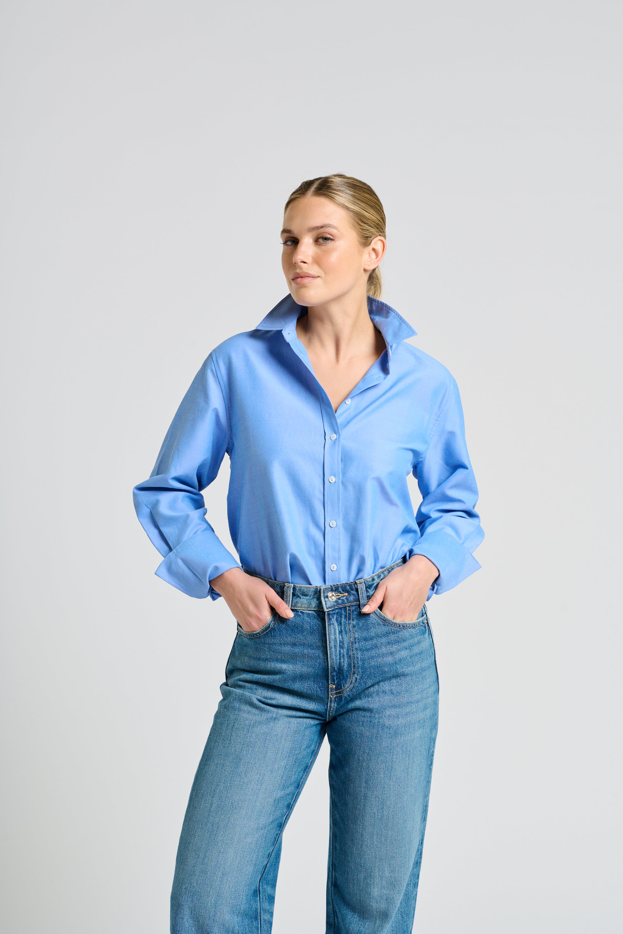 A woman modeling a light blue long-sleeve button-up shirt tucked into blue jeans, standing against a solid light gray studio background.