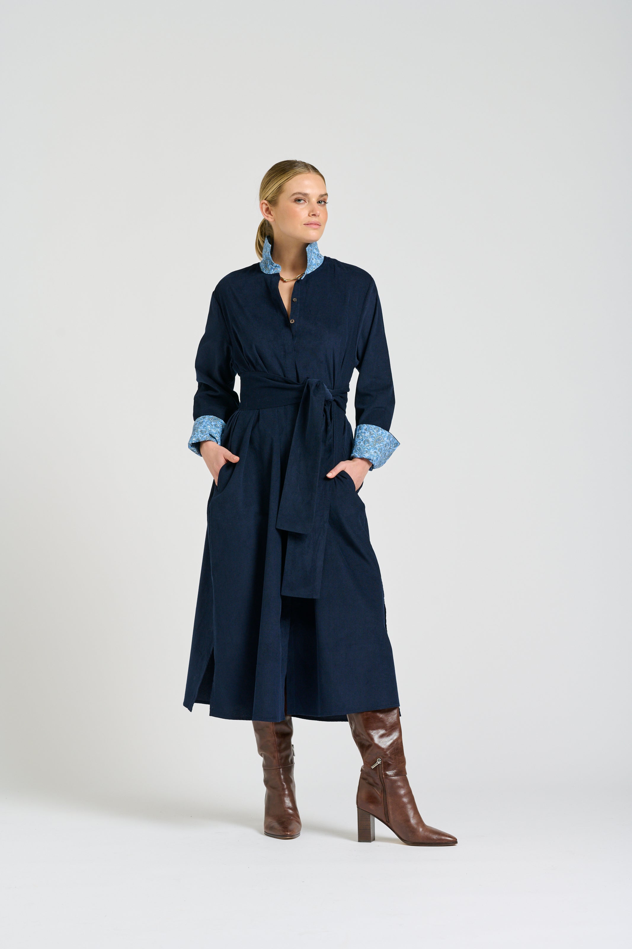 A woman models a navy blue wrap-style coat with light blue patterned lining on the collar and cuffs, paired with brown knee-high boots, against a plain light gray studio background.