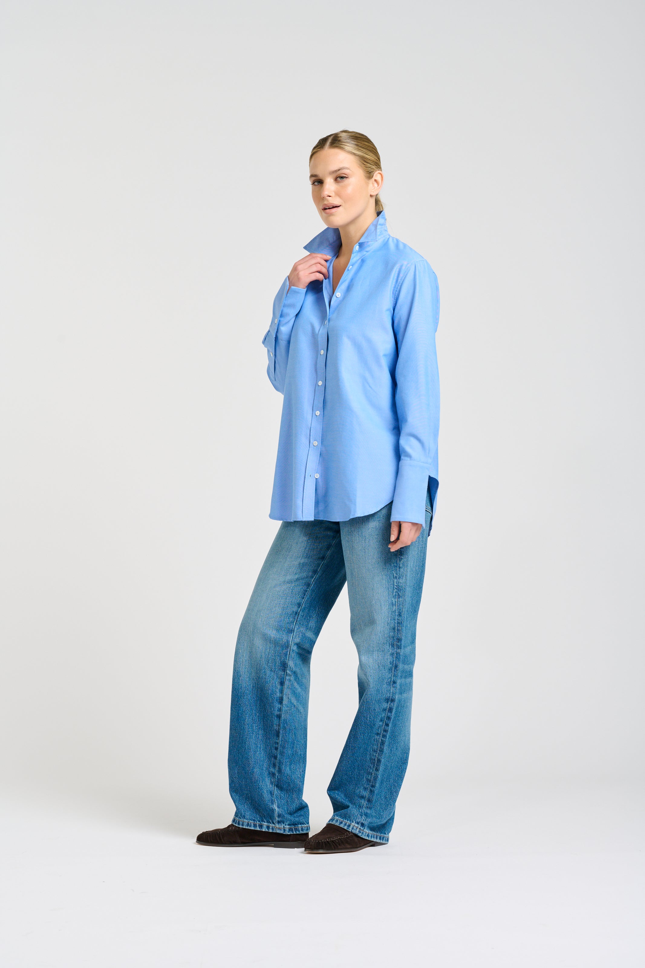 A woman modeling a light blue long-sleeve button-up shirt and wide-leg blue jeans against a plain light gray studio background.