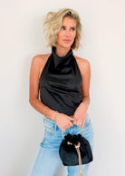 A woman wearing a black satin halter top and light blue jeans, holding a small black velvet drawstring bag with a gold chain strap against a plain white background.