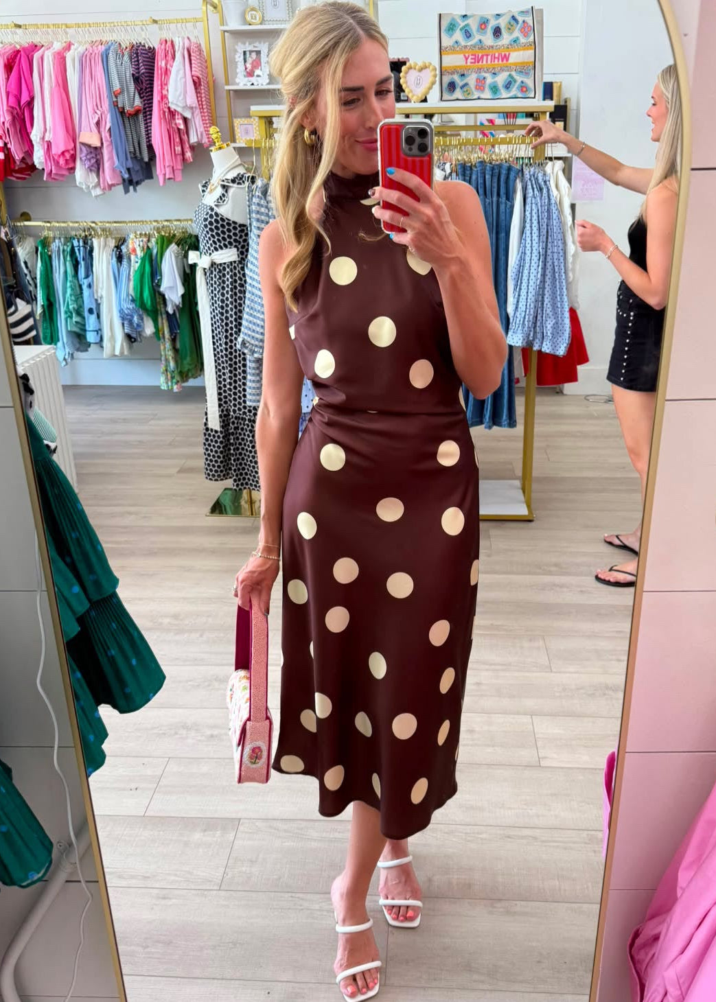A woman takes a mirror selfie in a clothing boutique wearing a brown sleeveless midi dress with large beige polka dots. The boutique has pink walls, a large arched mirror, and racks of colorful children's clothing in the background.