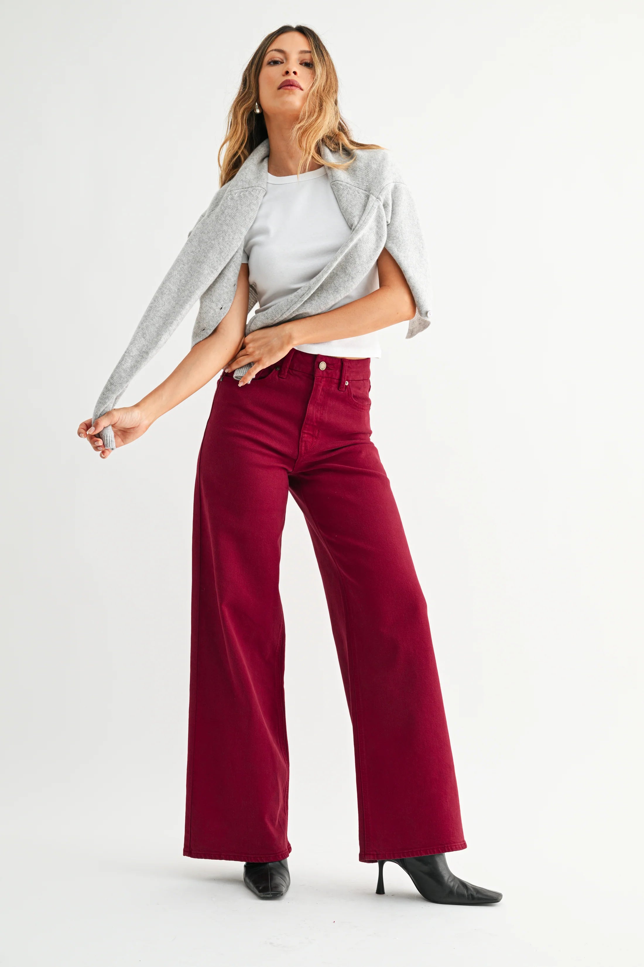 A woman modeling a pair of wide-leg trousers in deep burgundy, styled with a white top, a light gray cardigan, and black pointed-toe heels against a plain white studio background.
