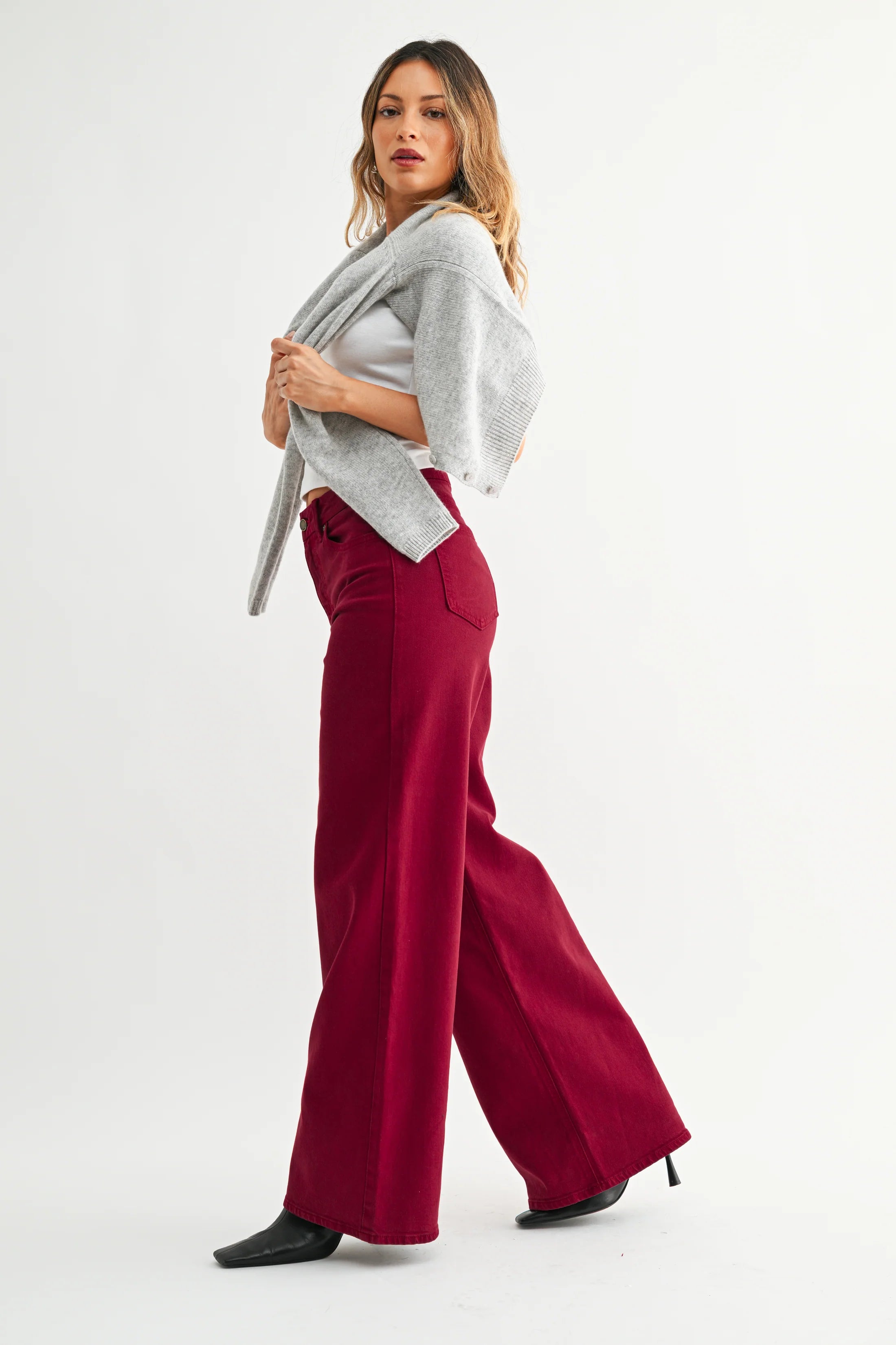 A woman modeling a pair of wide-leg burgundy trousers, styled with a light gray knit cardigan, a white top, and black pointed-toe heels, against a plain white studio background.