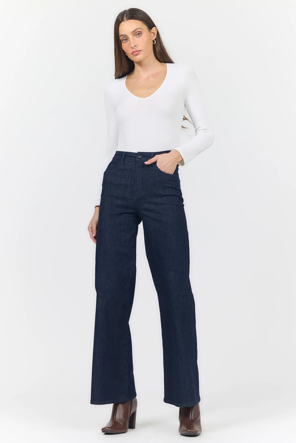 A woman modeling a pair of dark navy blue wide-leg jeans with a white long-sleeve top and brown ankle boots, standing against a plain white studio background.