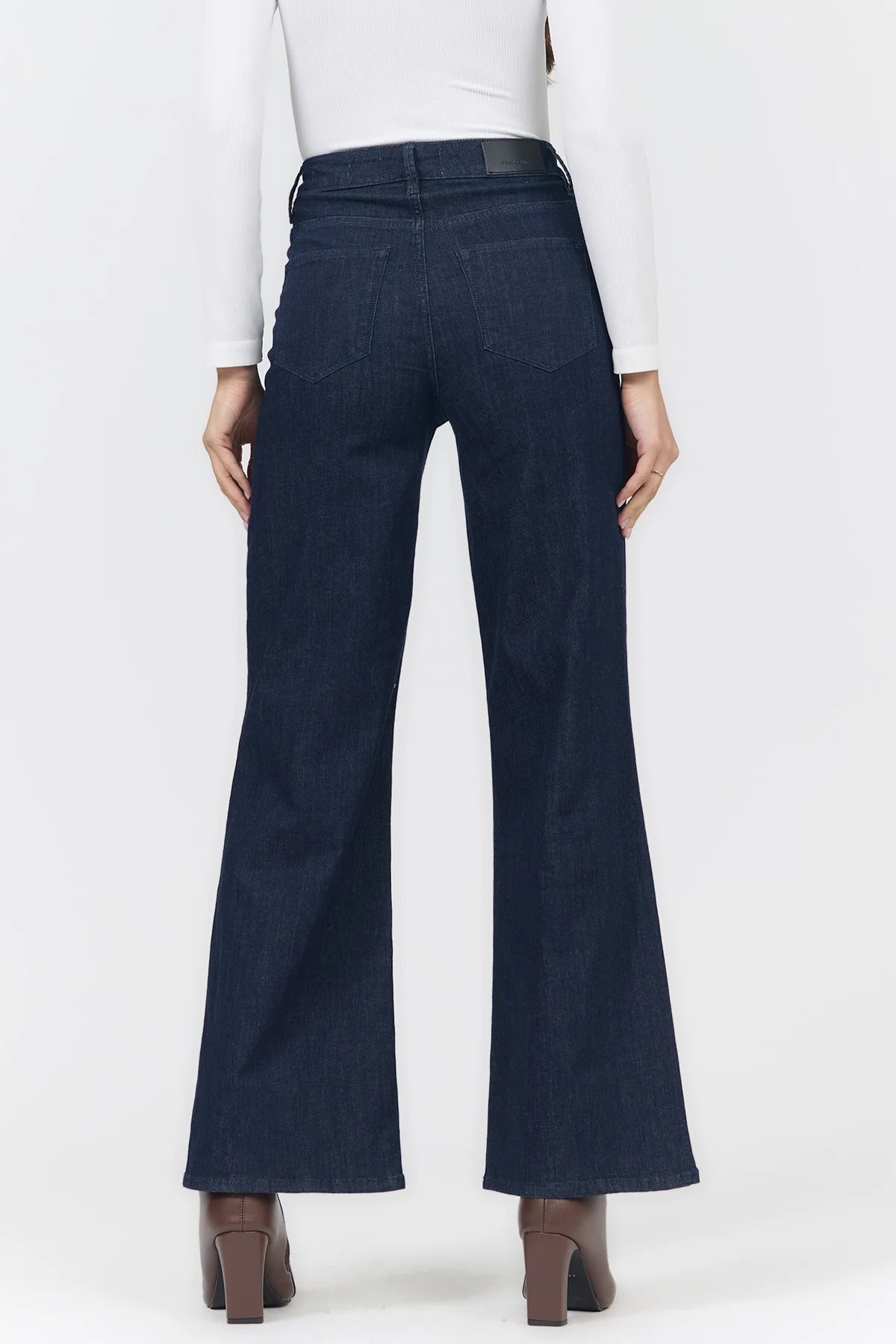Rear view of a person wearing dark blue wide-leg flare jeans with a white long-sleeve top and brown heeled shoes against a light gray studio background.