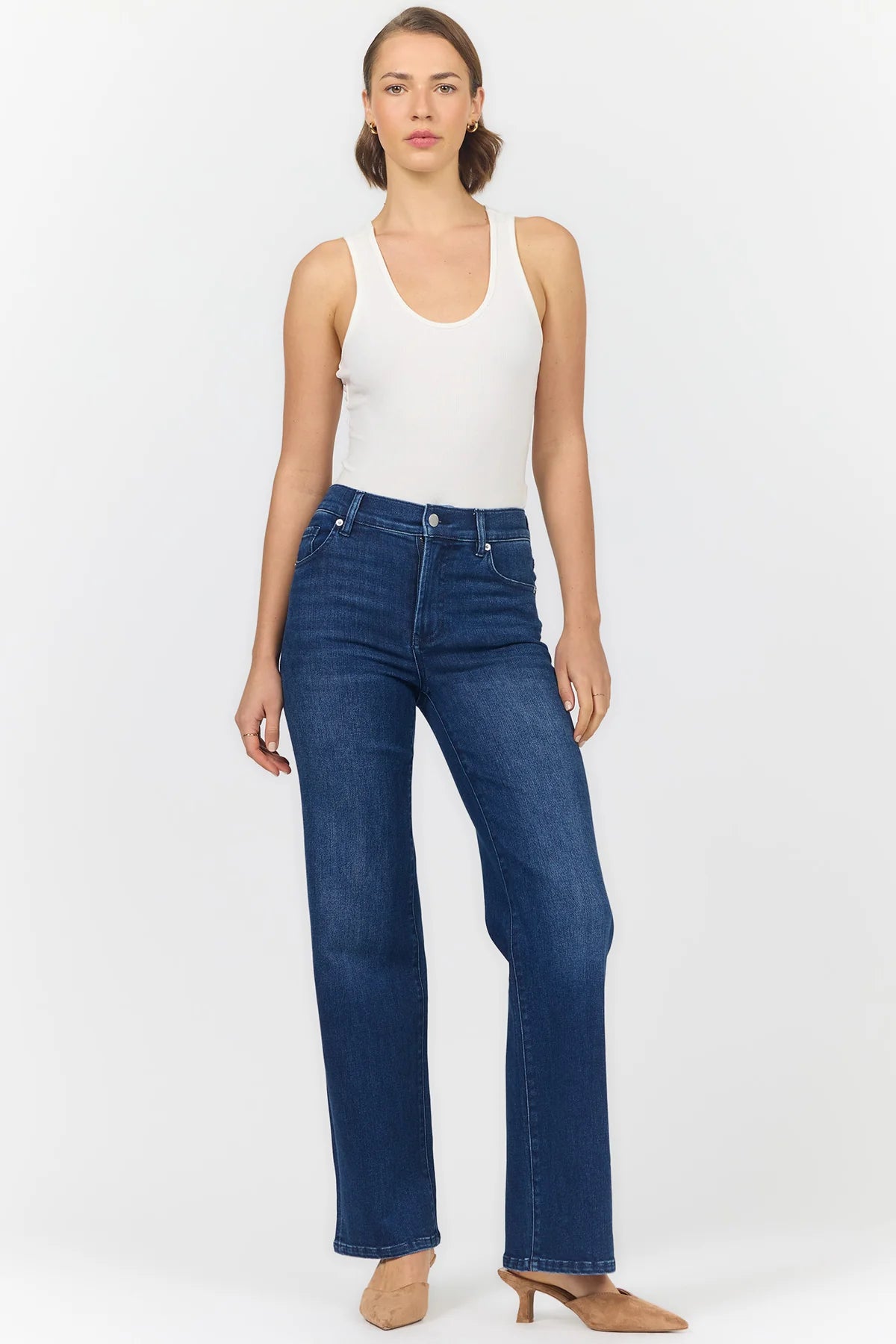 A woman modeling a pair of dark blue wide-leg jeans with a white tank top and beige heels, standing against a plain white studio background.
