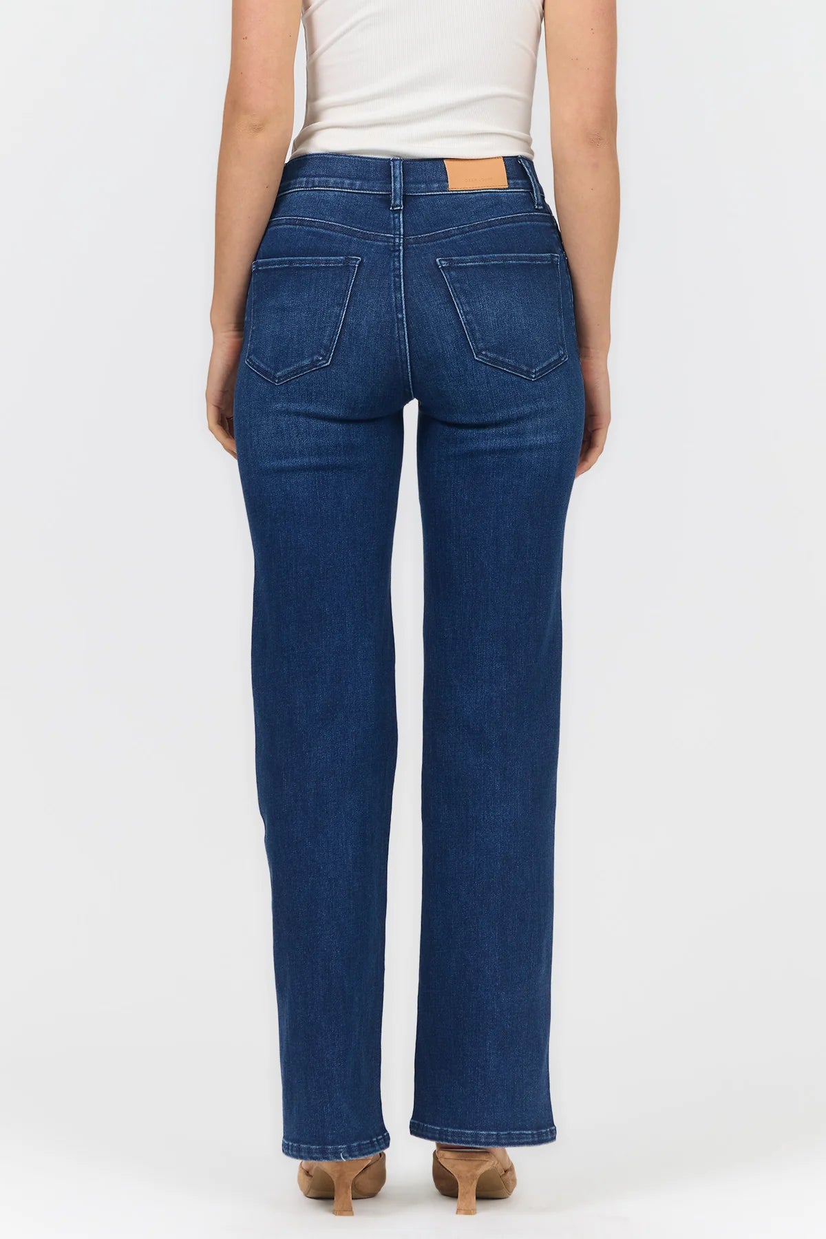 Rear view of a model wearing dark blue straight-leg jeans with a white top and tan heels, against a light gray studio background.