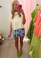 A woman takes a mirror selfie in a boutique showing off a cream short-sleeve button-up top and light blue shorts with dark blue polka dots. She is wearing bright green clogs, with clothing racks and a pink curtain visible in the background.