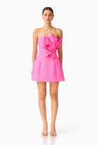 A model wearing a bright pink strapless mini dress with a large sculptural bow detail on the bodice, photographed against a clean white studio background.