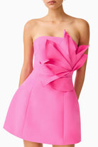 A bright pink strapless mini dress with a large sculptural layered bow detail on the bodice, shown on a model against a plain white background.