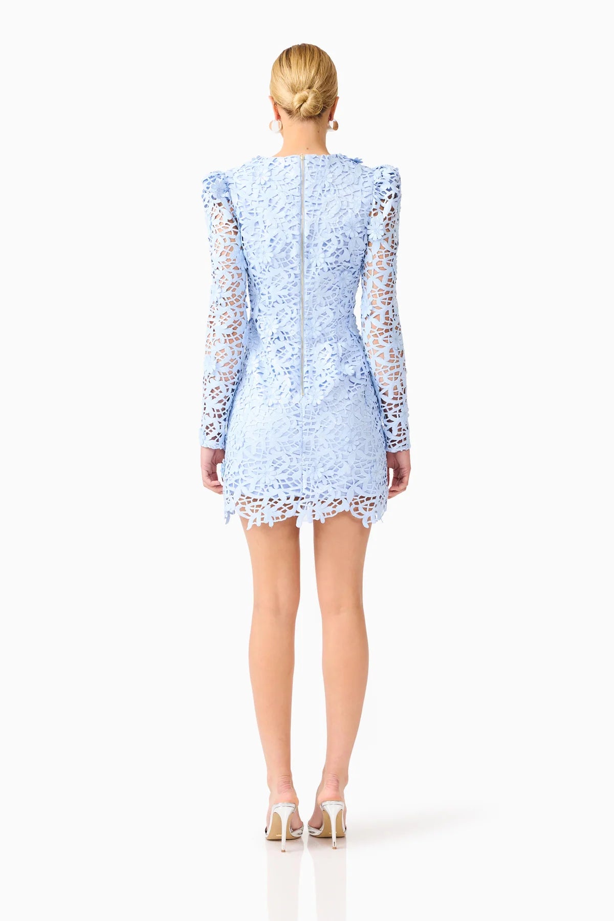 A woman viewed from the back wearing a light blue lace mini dress with long sheer lace sleeves and a scalloped hem, paired with beige high heels, against a plain white studio background.