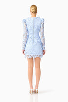 A woman viewed from the back wearing a light blue lace mini dress with long sheer lace sleeves and a scalloped hem, paired with beige high heels, against a plain white studio background.