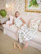 Woman in a floral dress sitting on a couch in a decorated room with floral wallpaper.