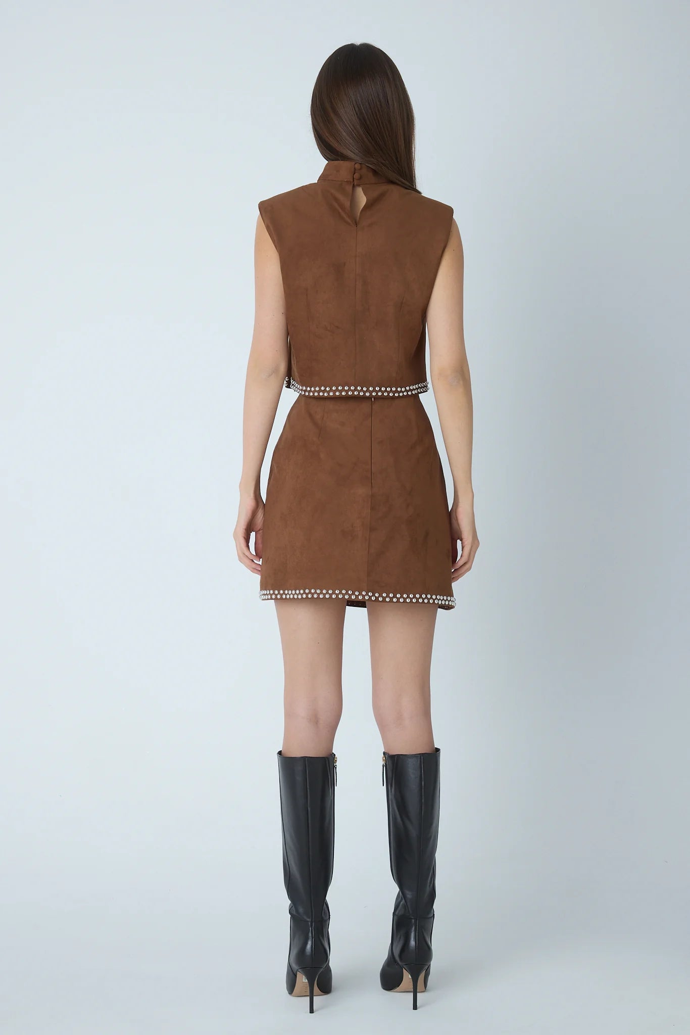 A woman shown from the back wearing a sleeveless brown mini dress with a high neck, a thin patterned waistband, and a matching patterned hem, paired with black knee-high boots against a light gray studio background.