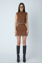 A model wearing a sleeveless brown faux-suede matching set with a mock neck and a thin black and white patterned waistband, paired with black knee-high boots against a light gray studio background.