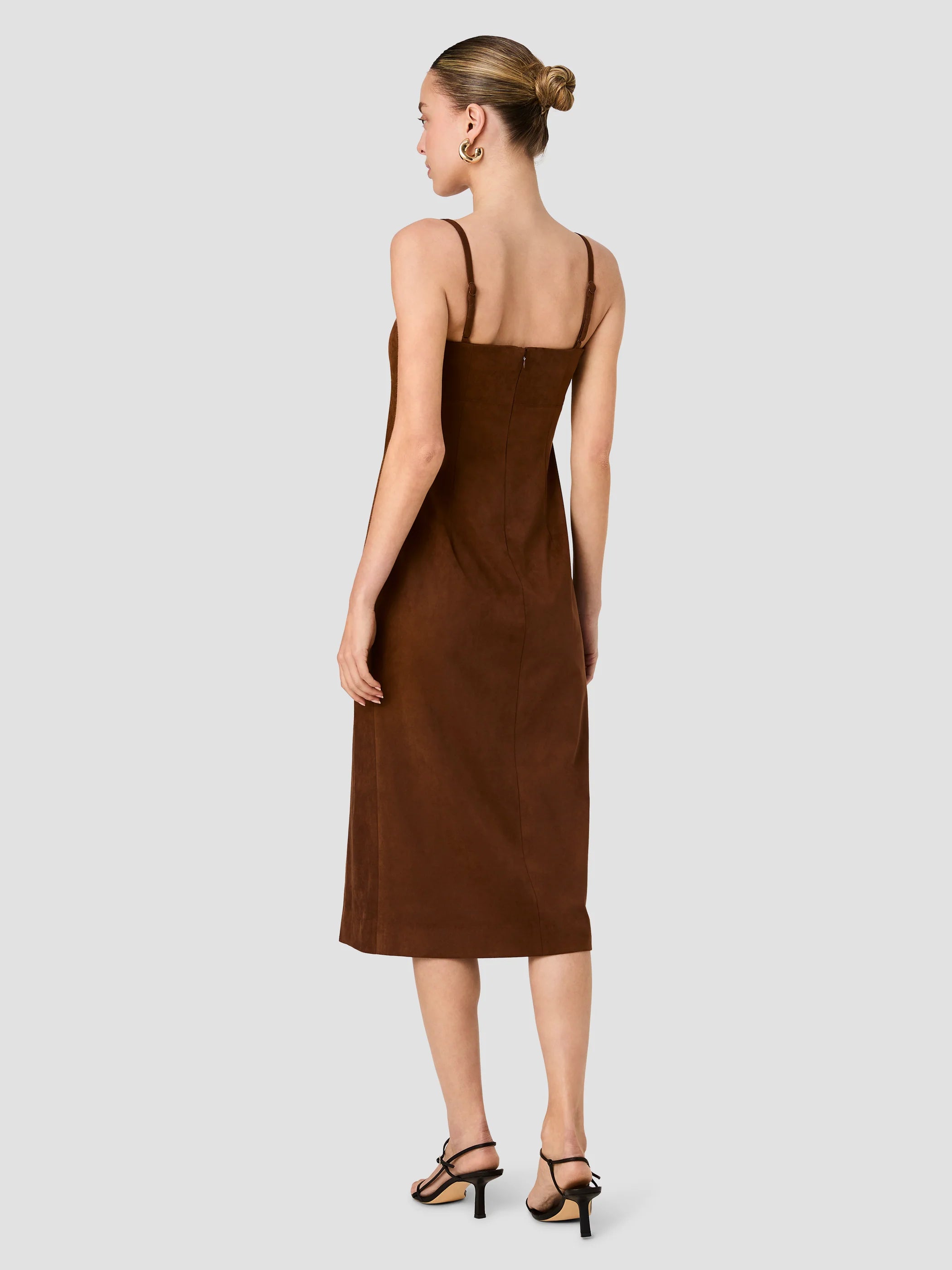 A woman shown from the back wearing a brown spaghetti-strap midi dress and black strappy heels against a light gray studio background.