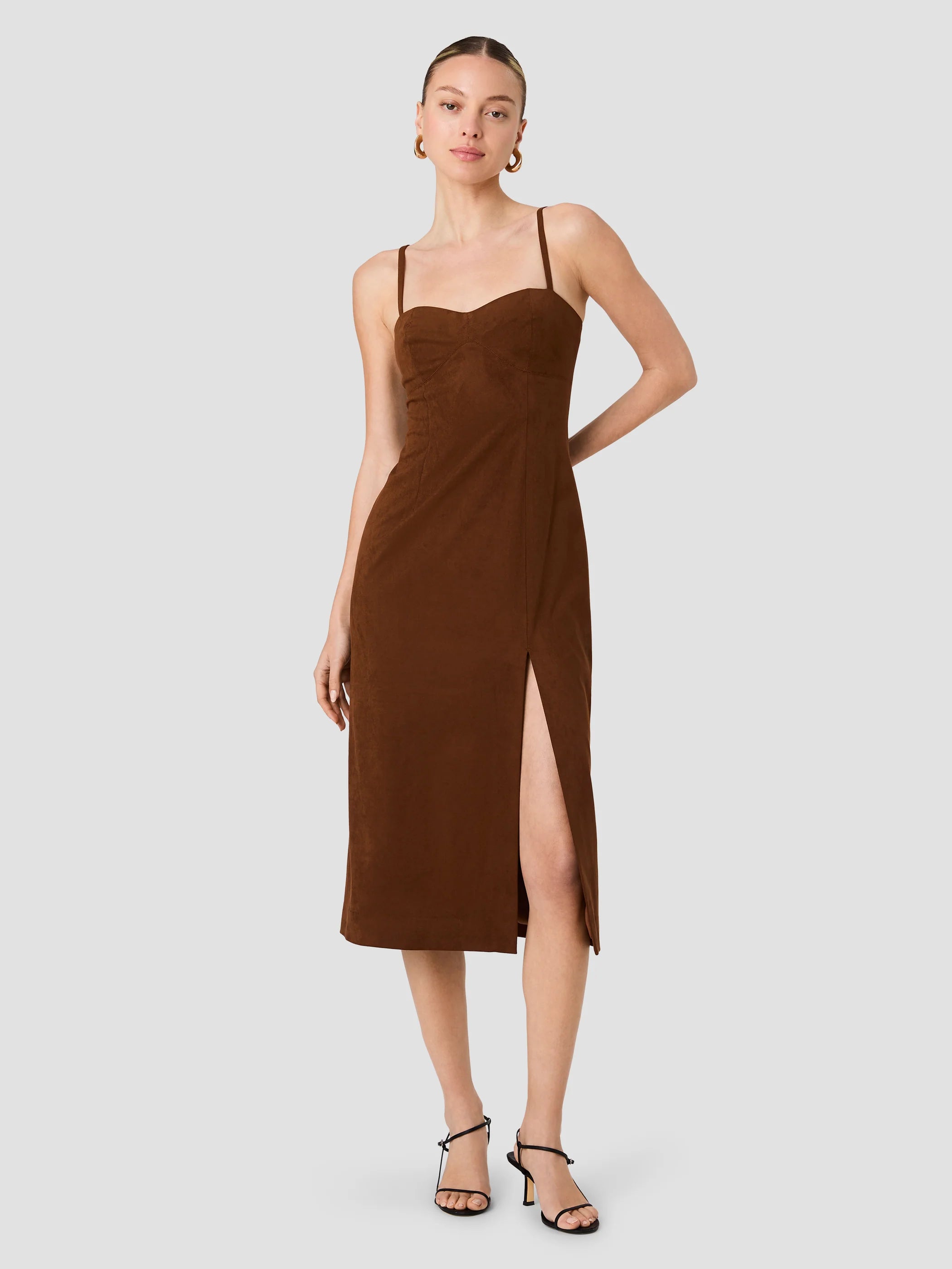 A woman modeling a brown midi dress with thin spaghetti straps, a sweetheart neckline, and a high side slit, paired with black strappy heels against a solid light gray background.
