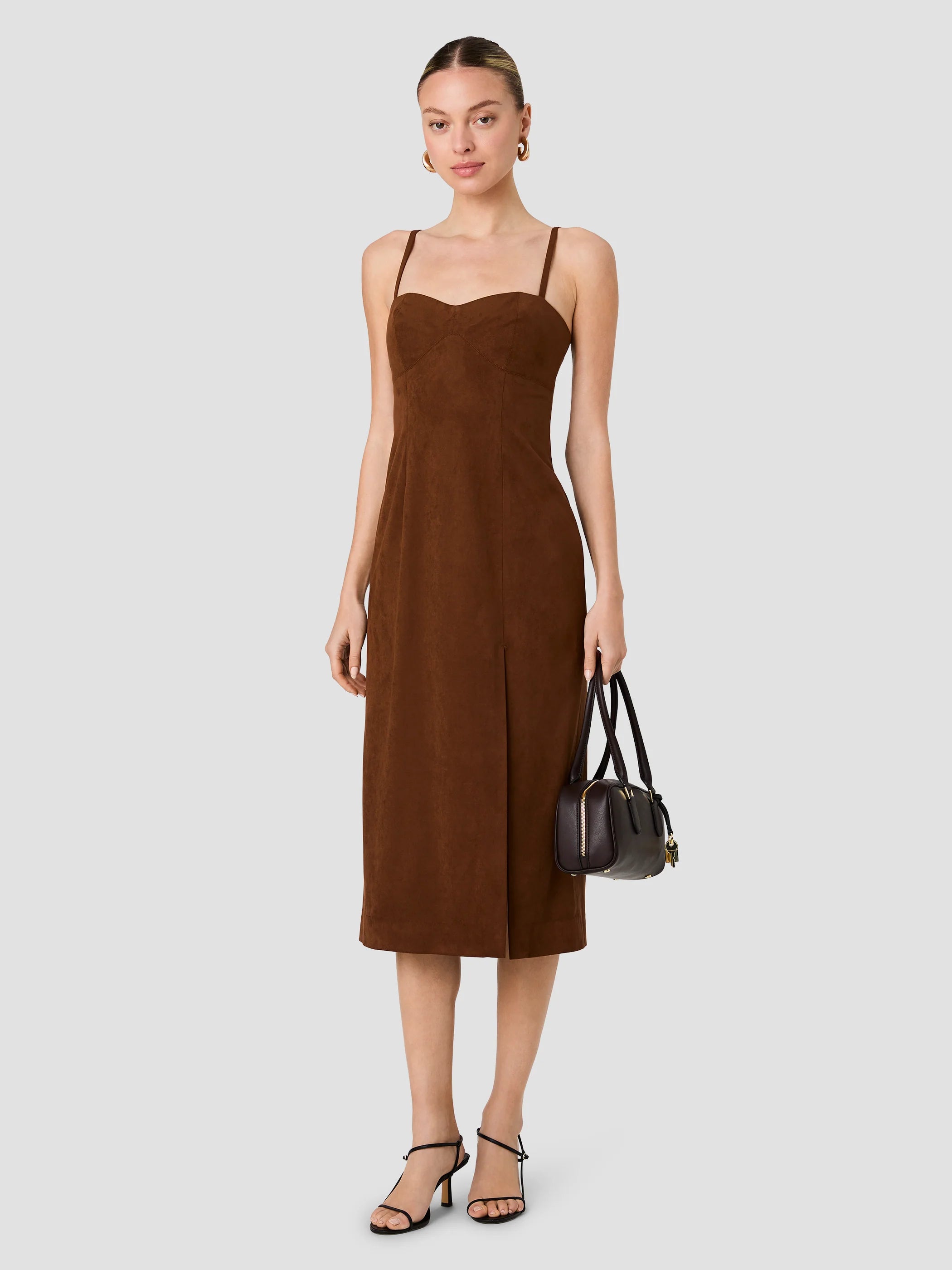 A woman modeling a brown sleeveless midi dress with thin straps, holding a small black handbag and wearing black strappy heels against a light gray studio background.