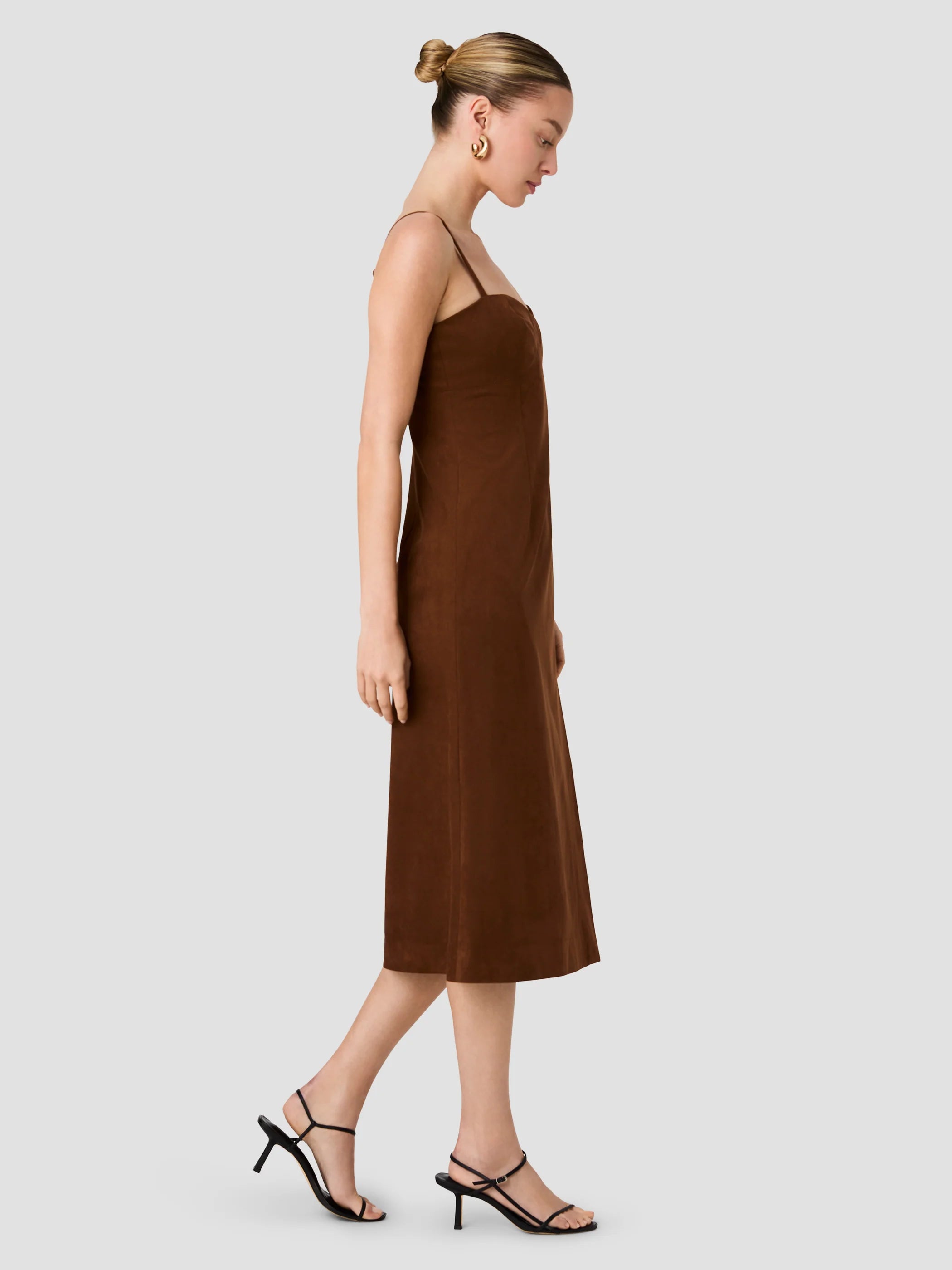 A woman modeling a brown midi-length spaghetti-strap dress in a side profile against a solid light gray background.