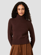A woman with curly dark hair wearing a dark brown ribbed mock neck sweater and a brown skirt, photographed against a solid light gray background.