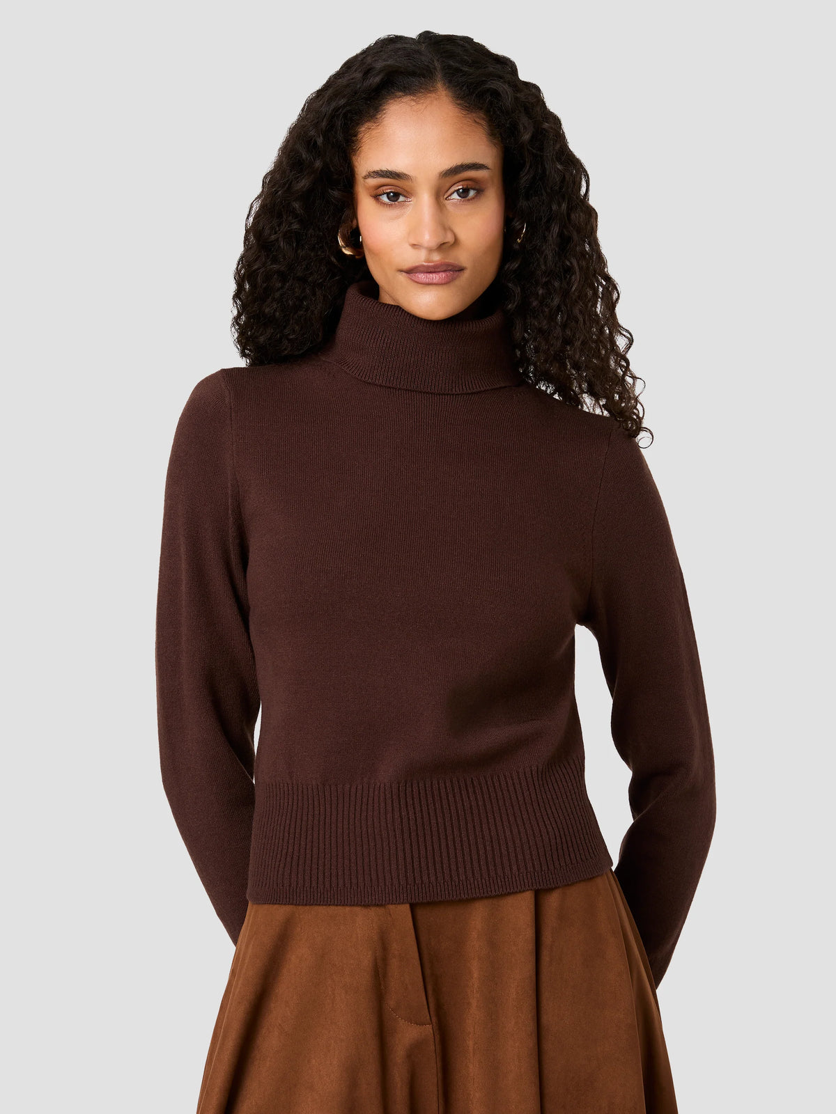 A woman with curly dark hair wearing a dark brown ribbed mock neck sweater and a brown skirt, photographed against a solid light gray background.