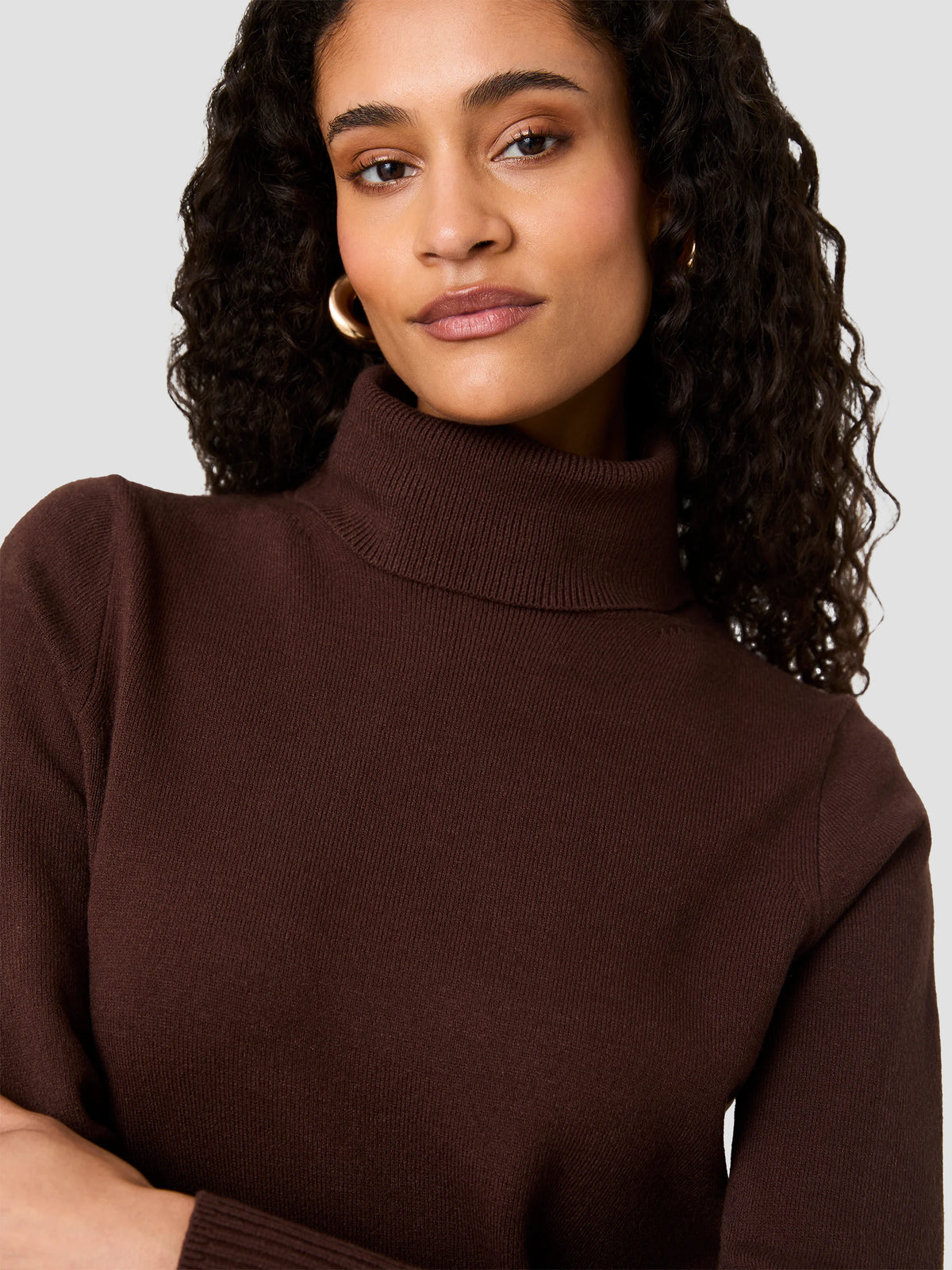 A close-up shot of a woman wearing a dark brown ribbed turtleneck sweater against a light gray background.
