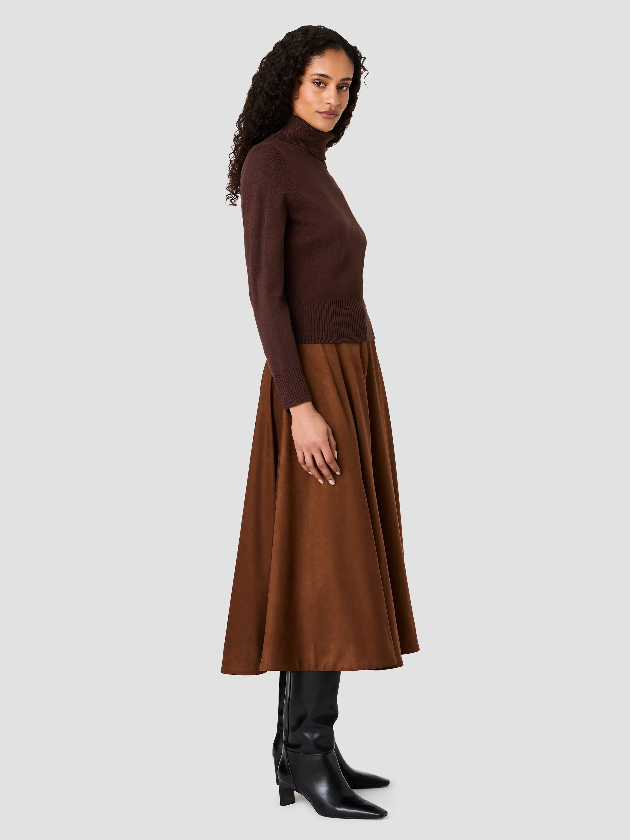 A woman modeling a dark brown mock-neck long-sleeve knit top paired with a matching brown midi skirt and black pointed-toe heeled boots, shown against a solid light gray background.