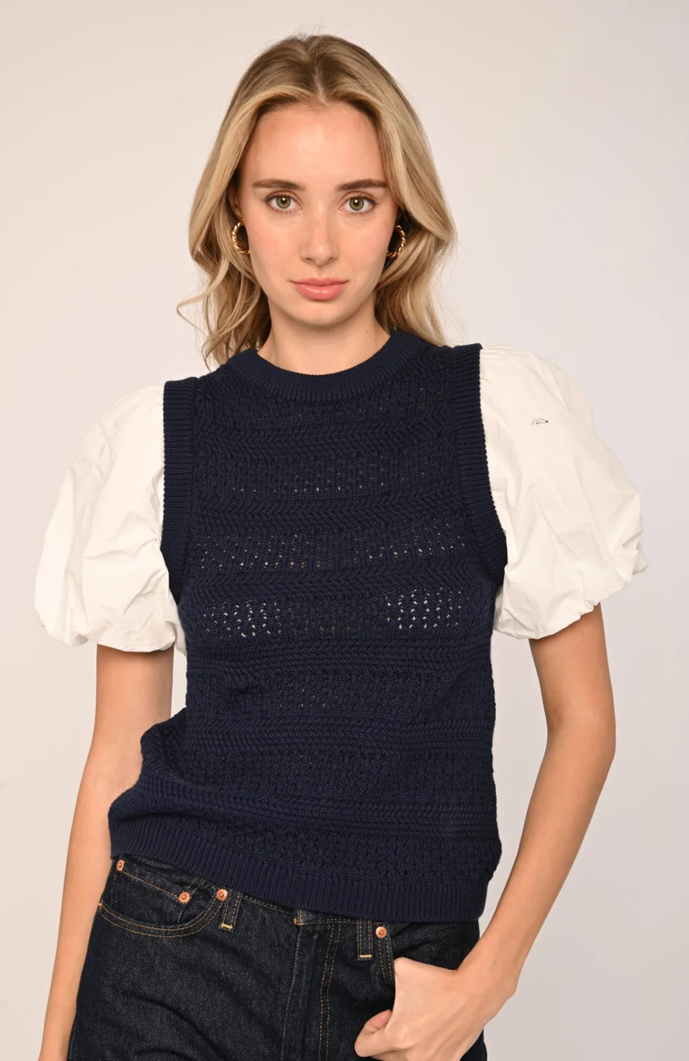 A woman modeling a sleeveless navy blue knit vest layered over a white puff-sleeve top and dark denim jeans, set against a plain light gray studio background.