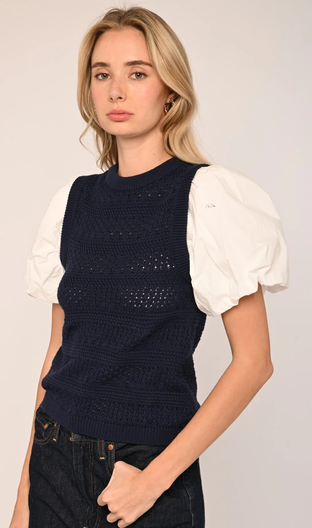 A woman modeling a sleeveless navy blue knit vest with textured openwork details over a white puff-sleeve top, paired with dark jeans, against a plain light gray studio background.