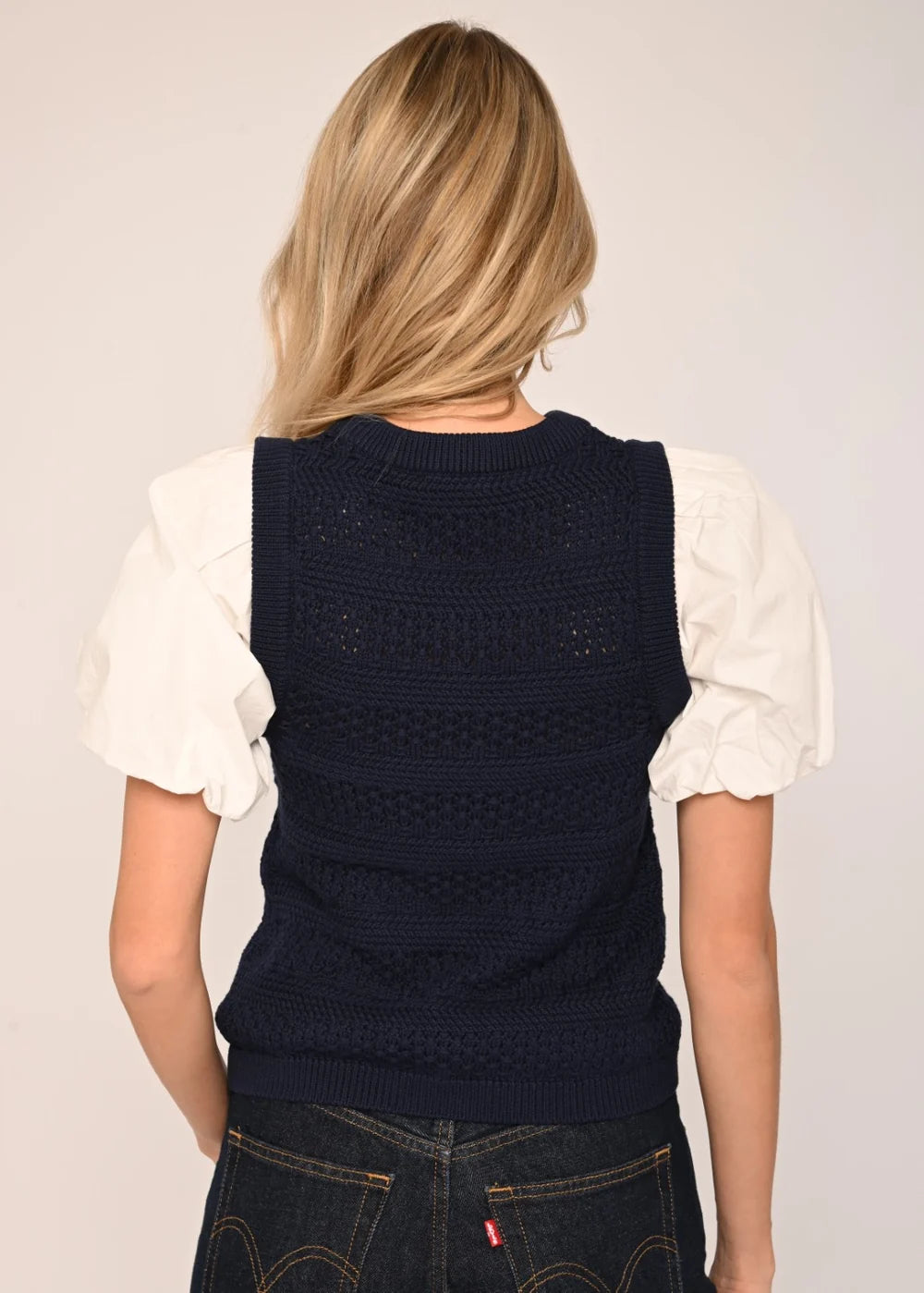A woman viewed from behind wearing a navy blue knit sleeveless vest over a white puff-sleeve blouse and dark blue jeans, against a plain light background.