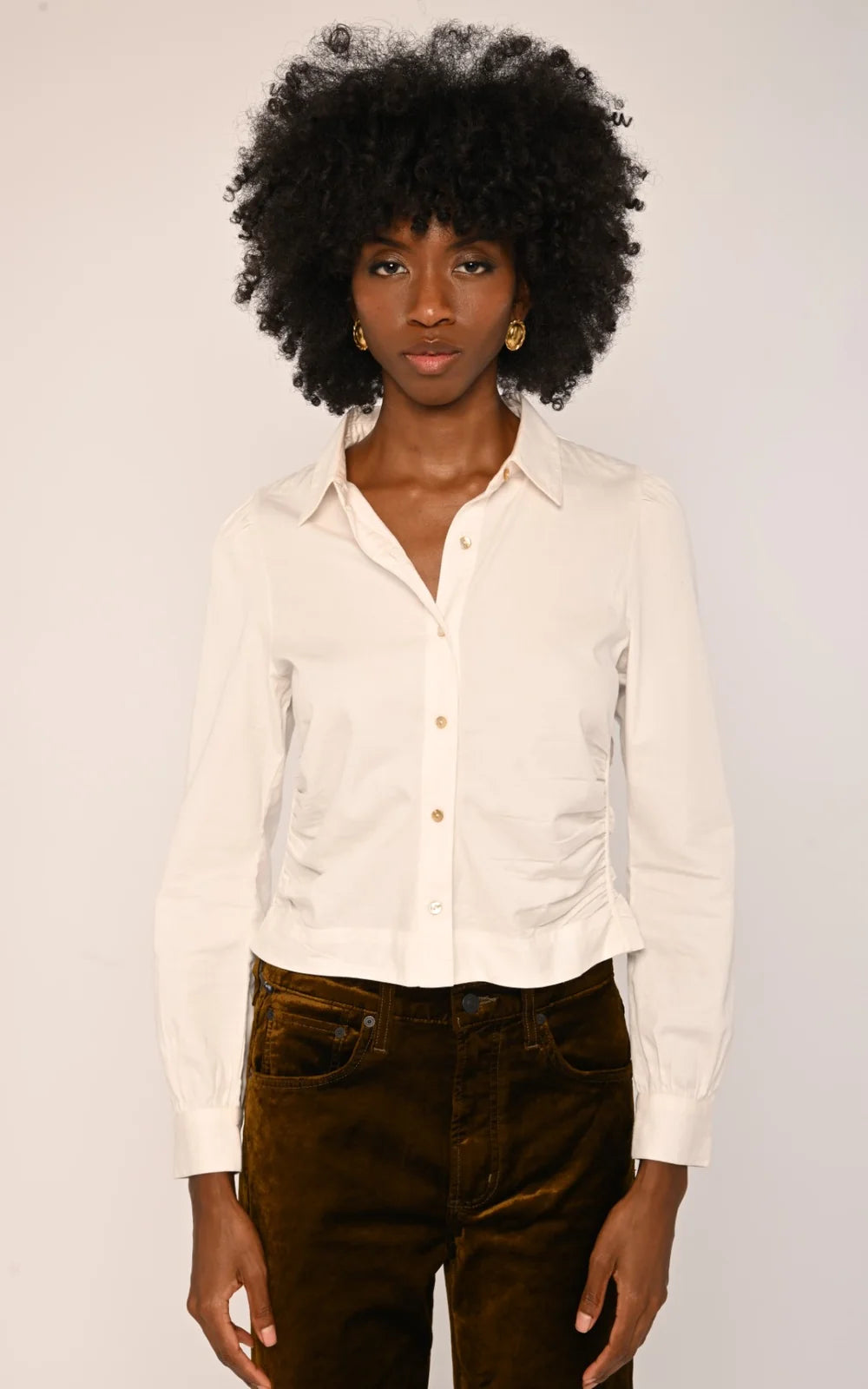 A woman with a large black afro hairstyle wearing a cream-colored long-sleeve button-up shirt and dark brown velvet pants, standing against a plain off-white background.