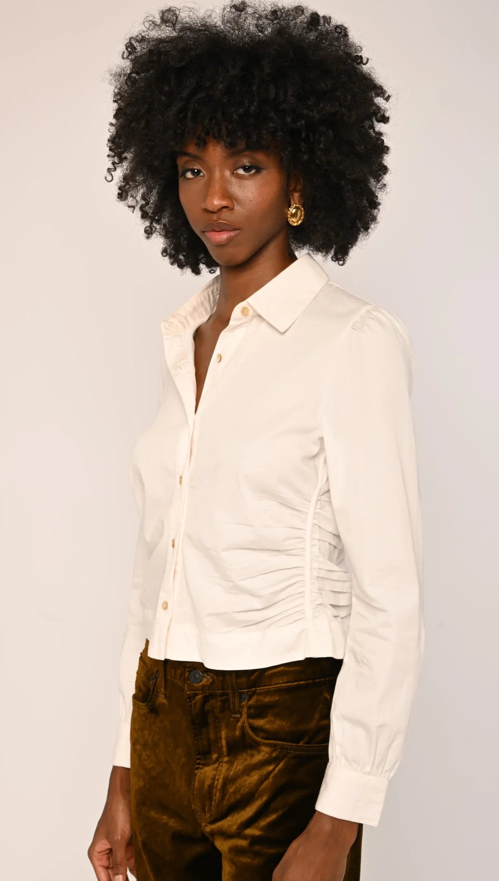A woman with a large black afro wearing a cream-colored long-sleeve button-up shirt with gathered detailing at the waist and brown velvet pants, photographed against a light gray studio background.