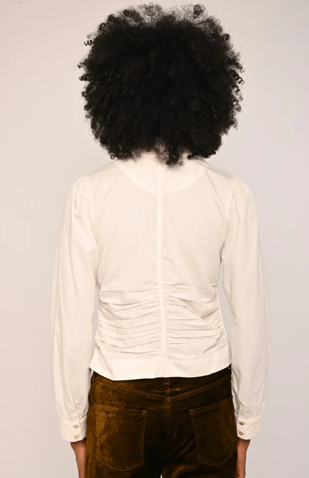 A person shown from the back wearing a fitted off-white long-sleeve blouse with gathered ruching at the back and dark brown velvet pants, against a plain light background.