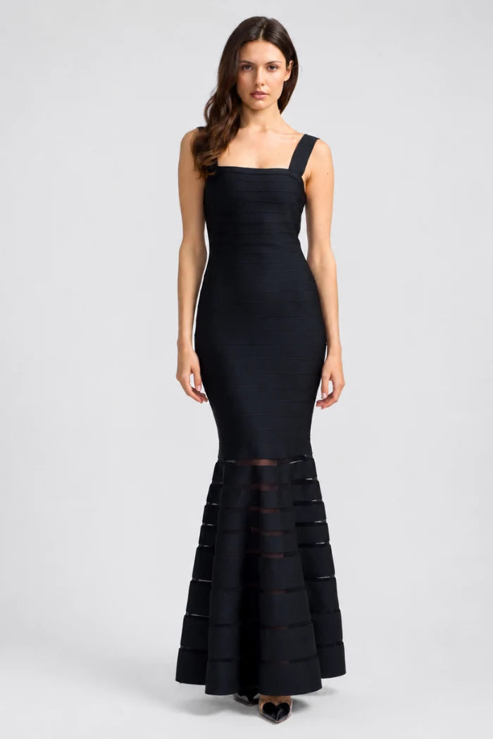 A woman modeling a black sleeveless mermaid gown with a square neckline, fitted bodice, and layered ruffle detailing at the flared hem, photographed against a light gray studio background.