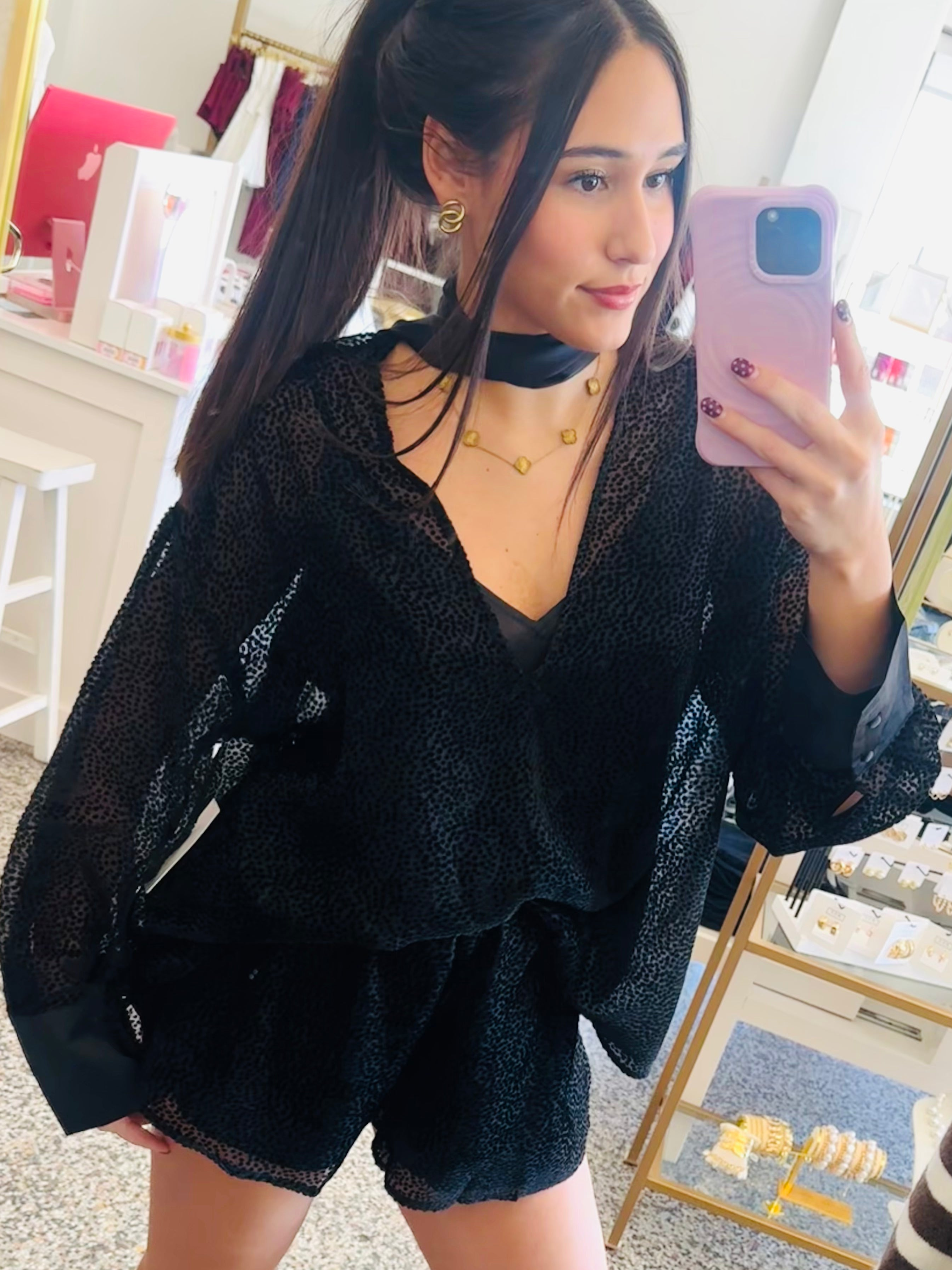 A woman takes a mirror selfie in a boutique, wearing a black sheer long-sleeve top and matching black shorts, paired with beige cowboy boots.