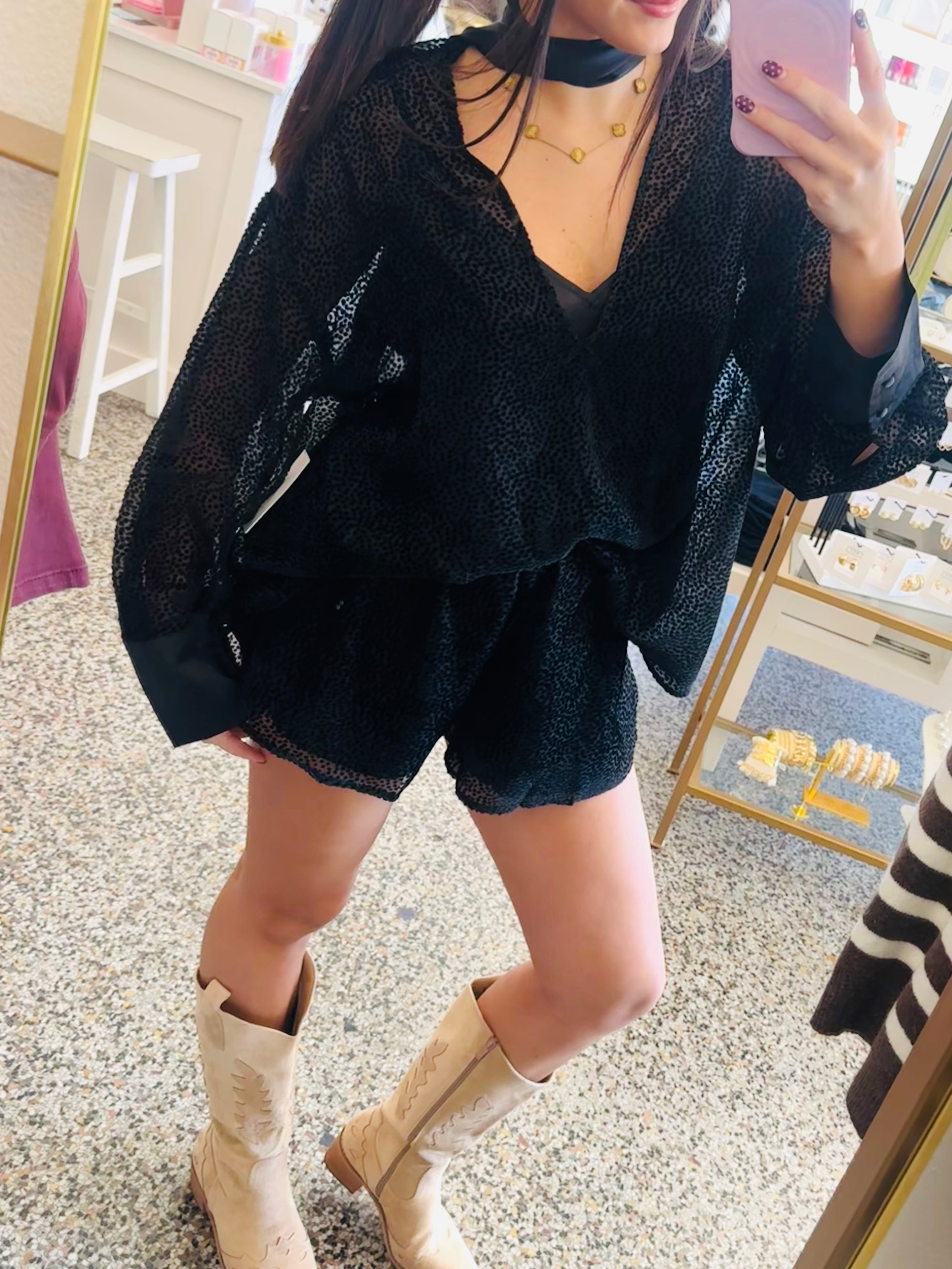 A woman takes a mirror selfie in a boutique, wearing a black sheer long-sleeve top and matching black shorts, paired with beige cowboy boots.