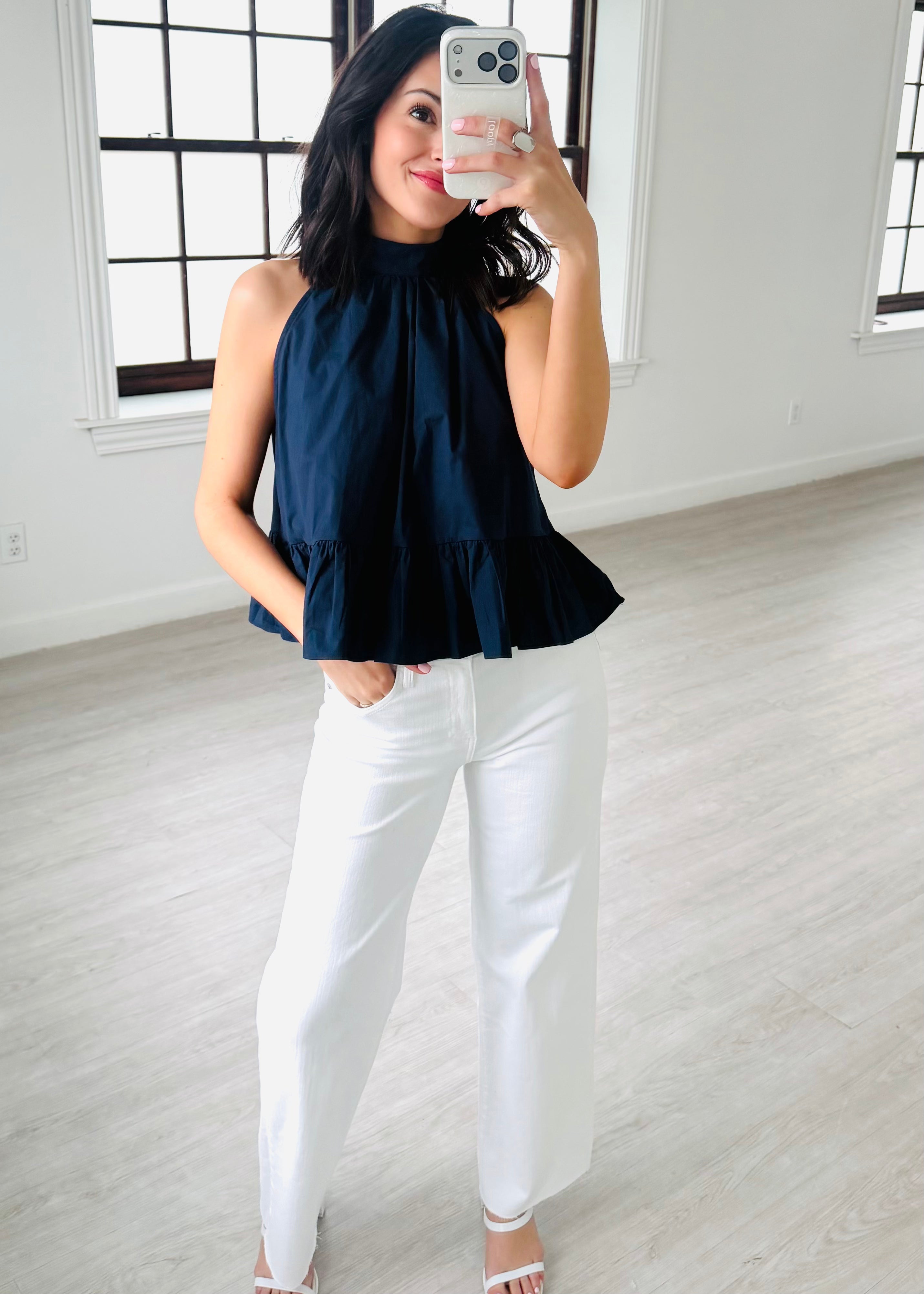 Woman taking a mirror selfie wearing a navy top and white pants in a bright room.