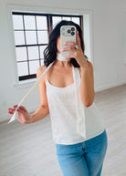 Person wearing a white tank top and blue jeans in a room with a window.