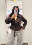 A woman takes a mirror selfie in a fitting room wearing a dark brown belted wrap jacket over a yellow top and leopard-print pants.