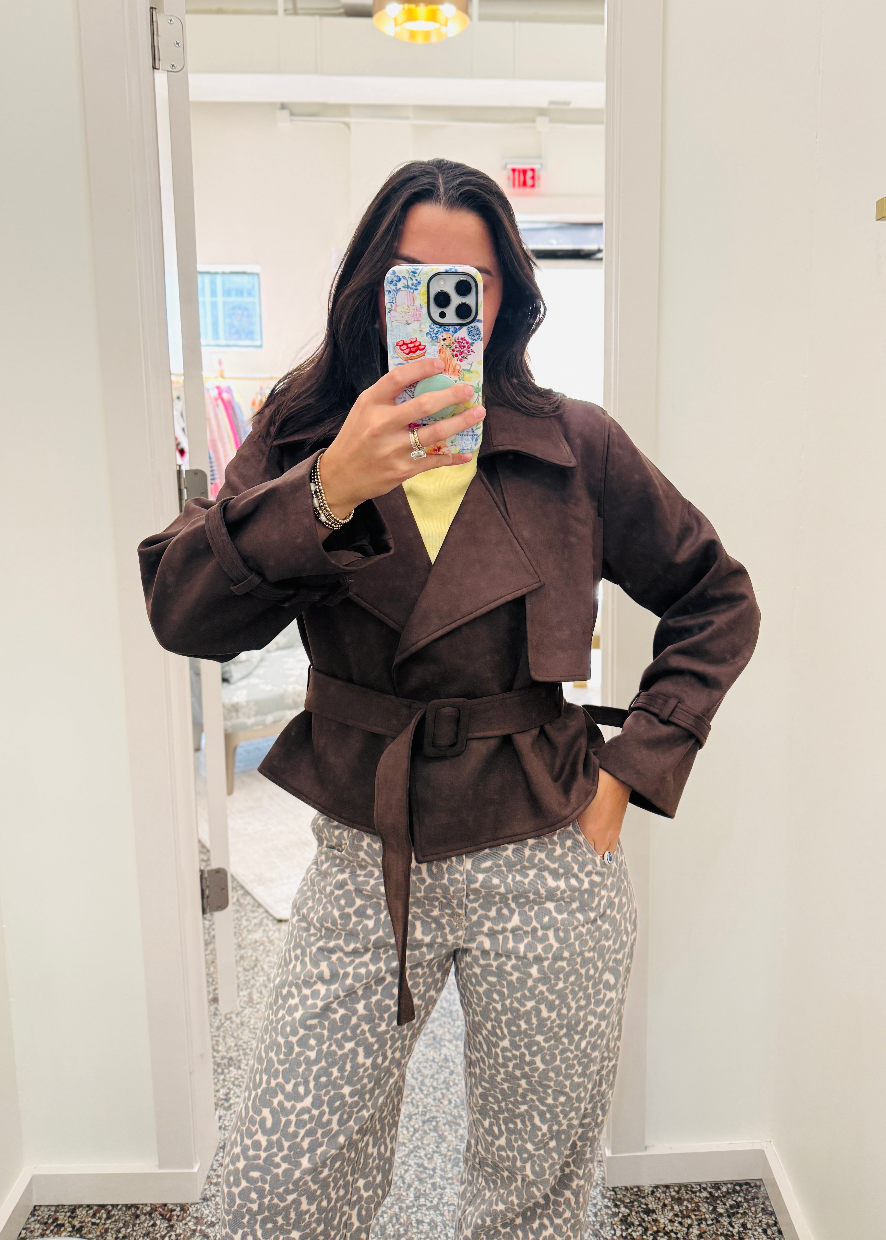 A woman takes a mirror selfie in a fitting room wearing a dark brown belted wrap jacket over a yellow top and leopard-print pants.