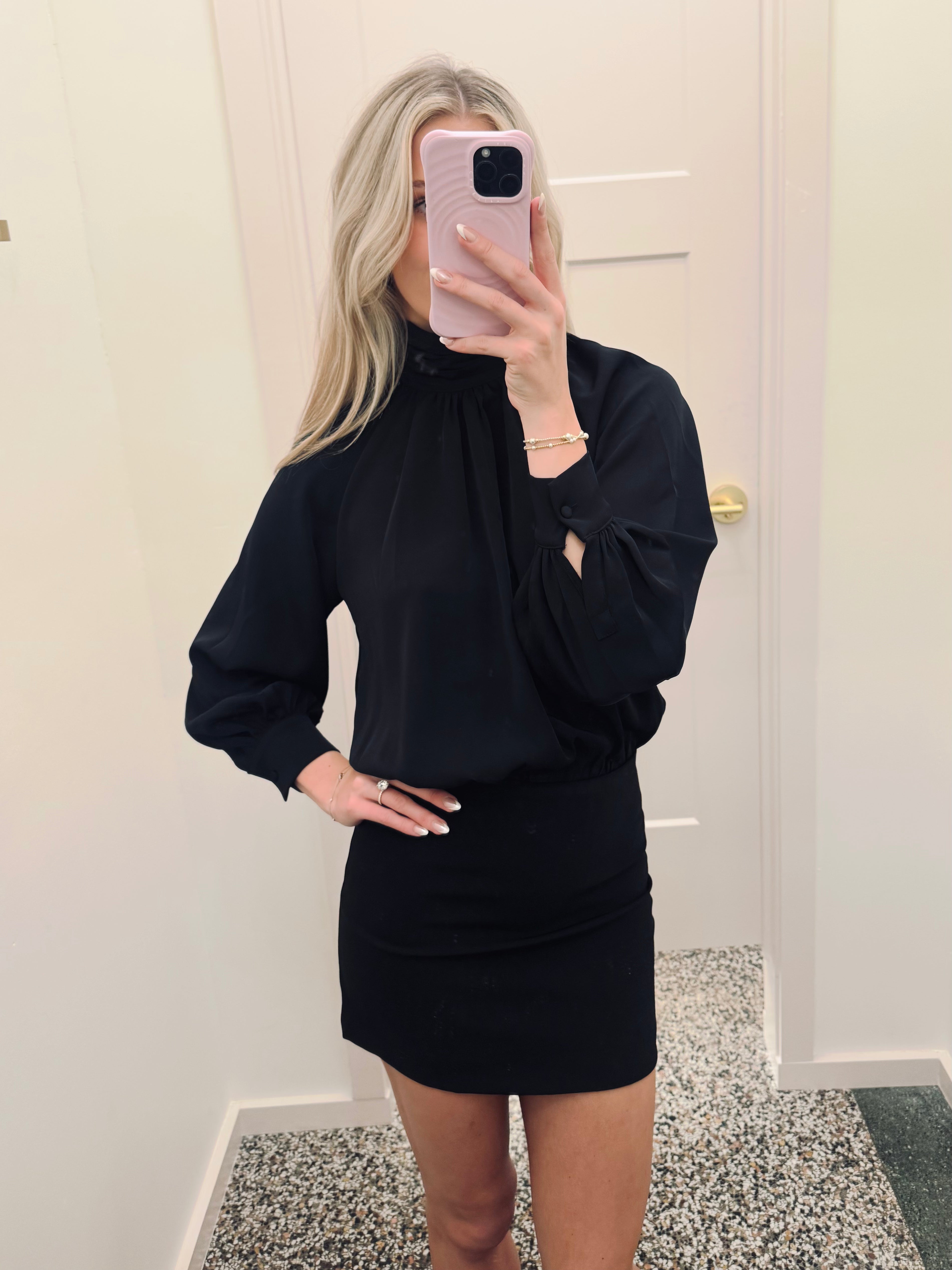 A woman takes a mirror selfie wearing a black long-sleeve mock-neck blouse with a high neck and a matching black bodycon mini skirt.